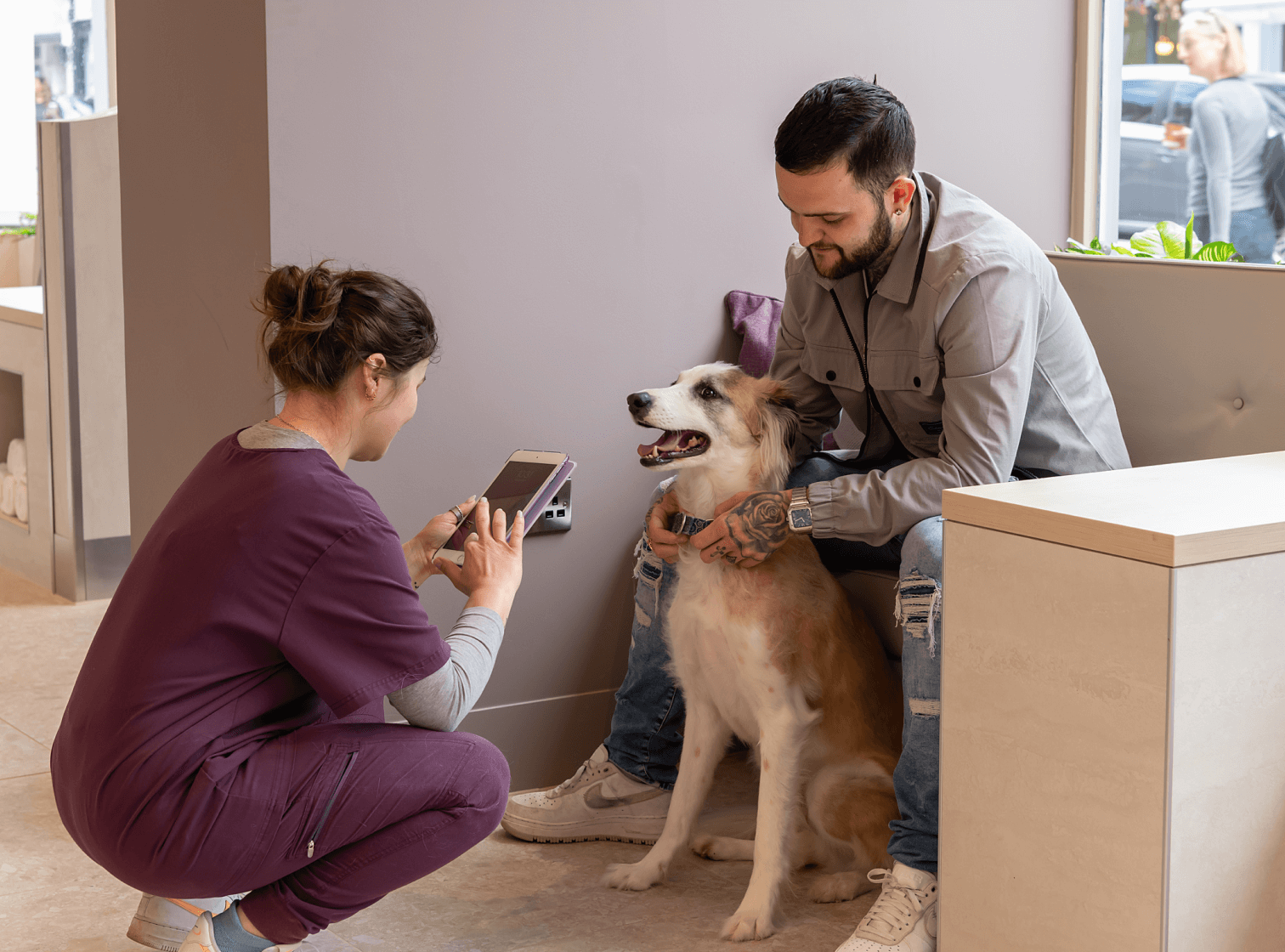 Person squatting to photograph a happy dog with another person in an indoor setting.