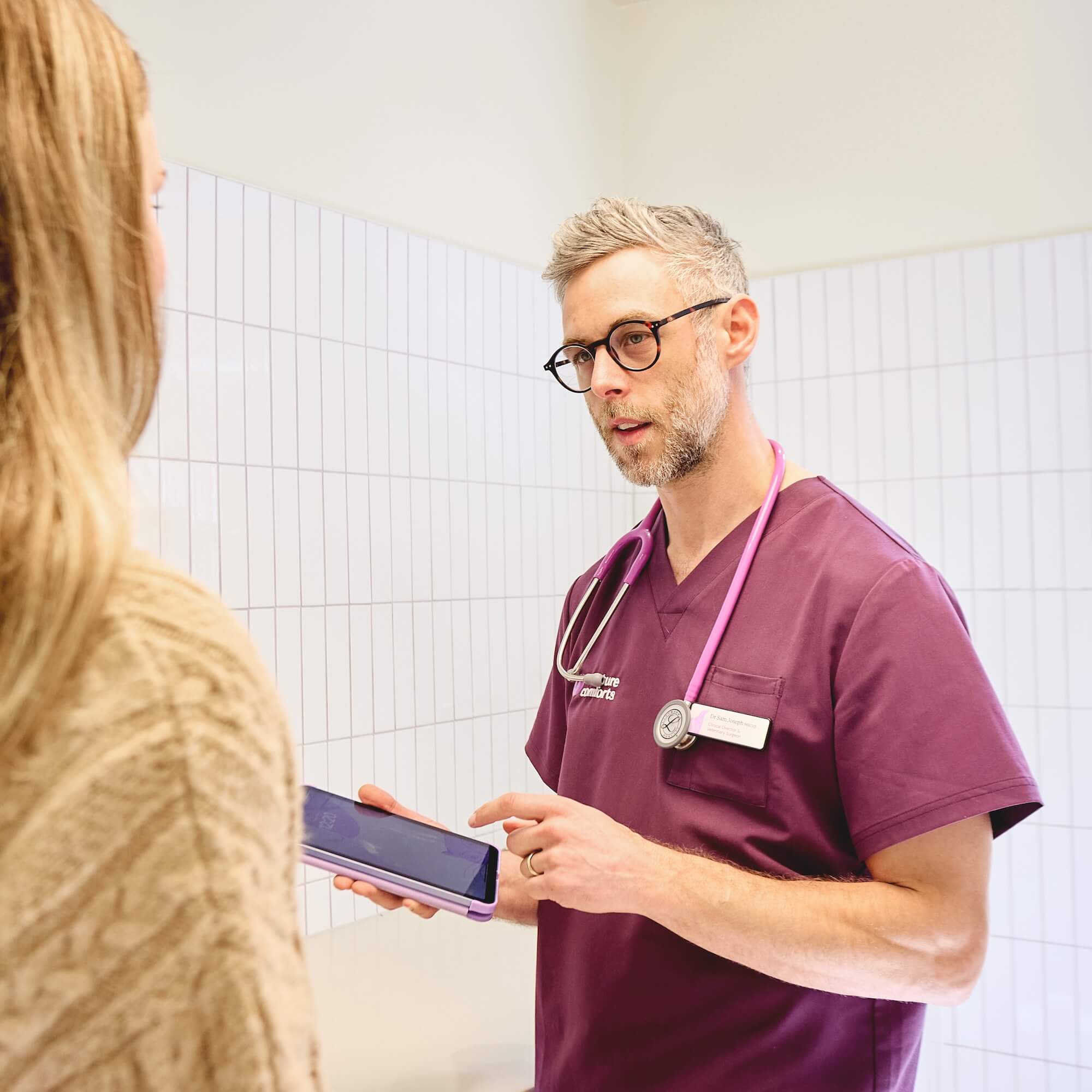 A healthcare professional in burgundy scrubs, with a stethoscope, engages a patient. They use a tablet in a well-lit, tiled room, conveying a focused, caring atmosphere.