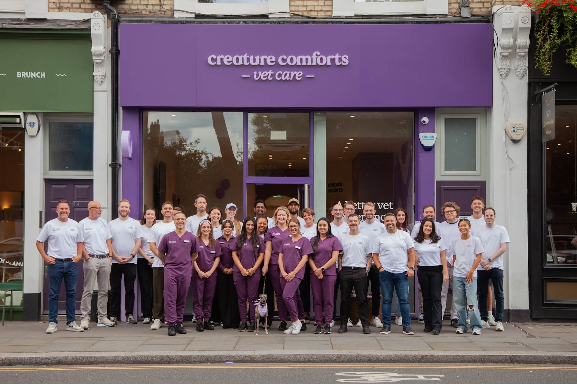 A group of smiling vet clinic staff stands outside under a purple storefront sign reading, "Creature Comforts Vet Care." They wear uniforms in white and purple.